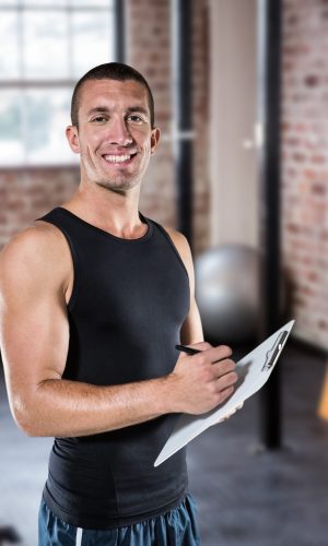 Smiling sports coach writing on clipboard against gym